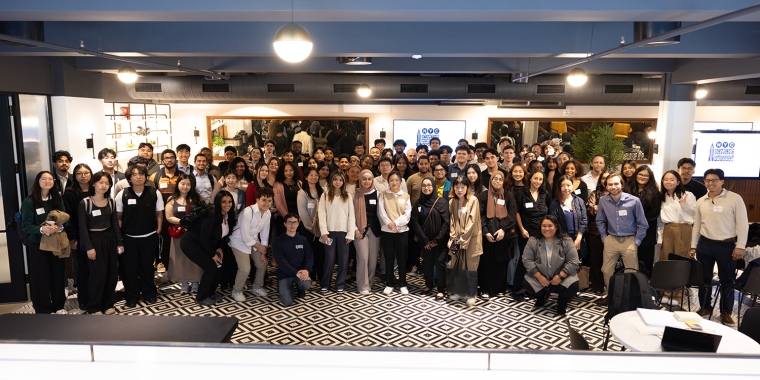NYC Startup Internship program participants pose for a group photo indoors, many wearing name tags and business attire, standing on a black-and-white patterned floor in a spacious, modern room.