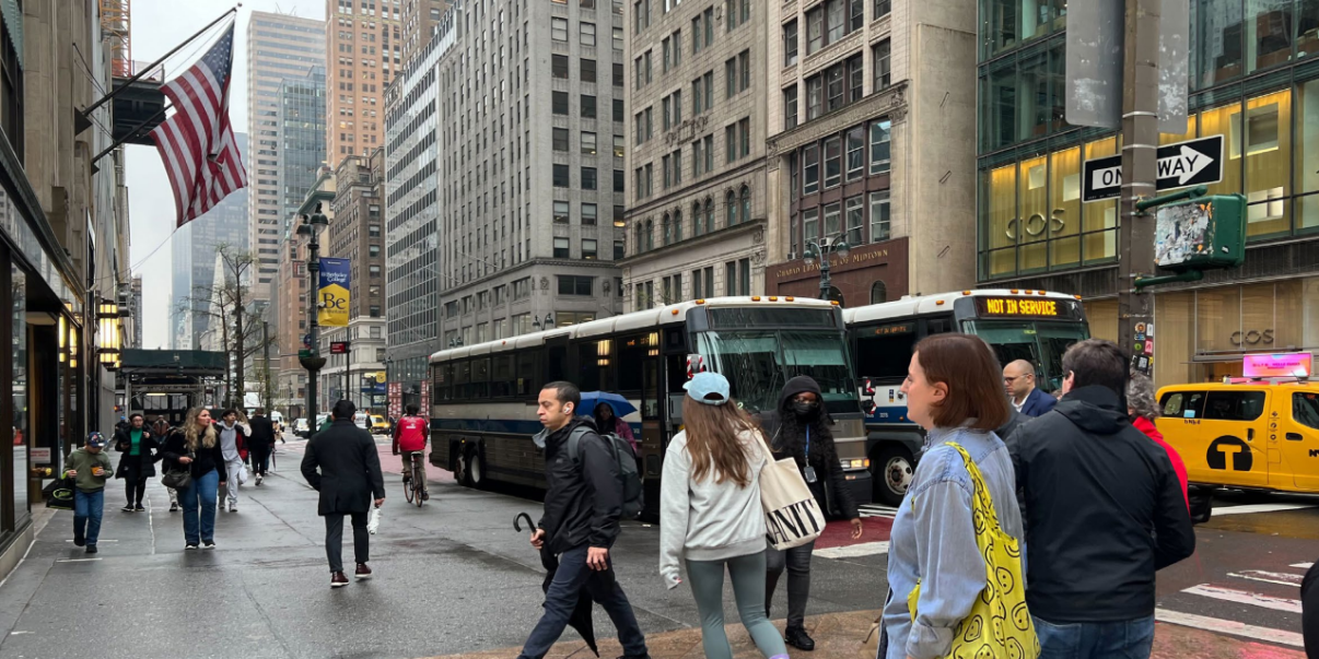 People walking on the busy 42nd Street in New York City with buses, taxis, and tall buildings in the background. An American flag is visible.