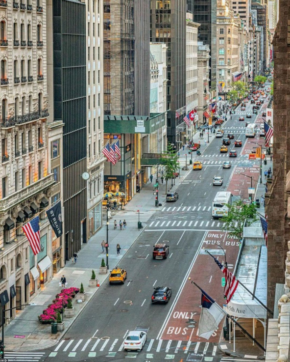 Aerial view of a busy Fifth Avenue in NYC with cars and buses, lined with tall buildings and American flags, featuring crosswalks and greenery along the sidewalks.